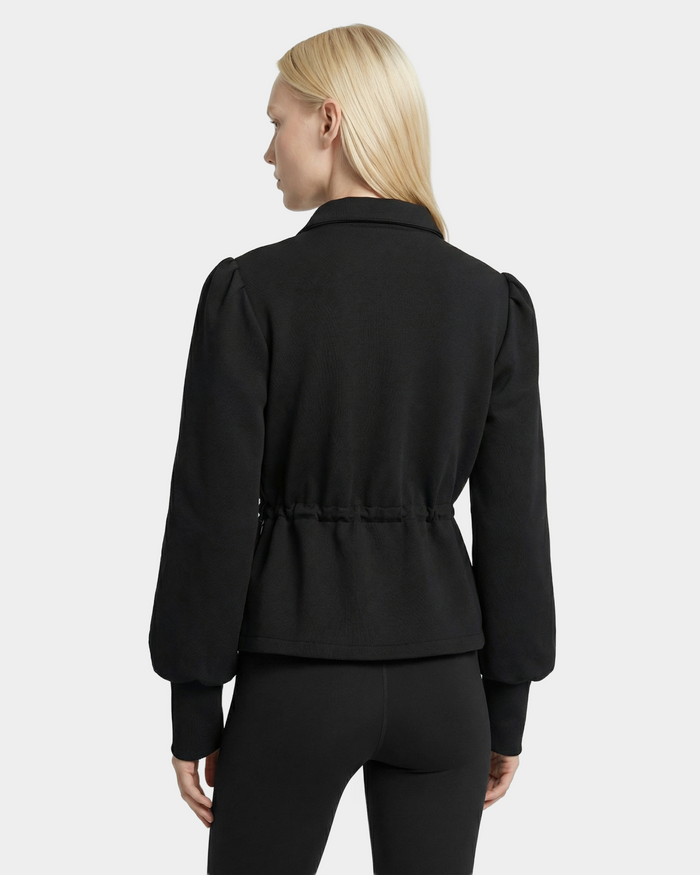 A person with straight blonde hair is shown from the back, wearing the Rudsak ELODIE - 8526039 Black zip-up sweater jacket with puffed long sleeves and fitted pants, standing against a plain light background.