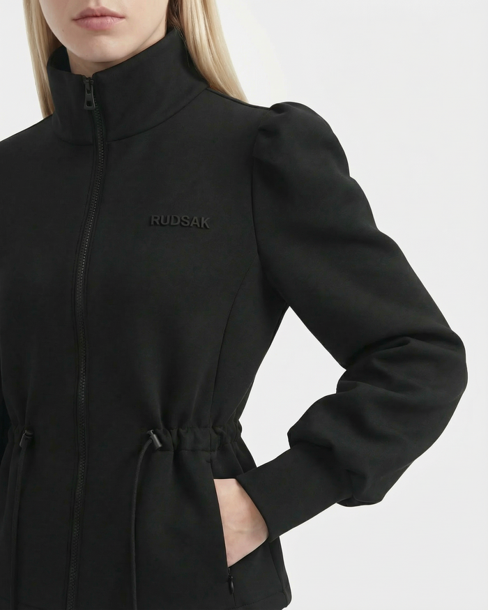 A woman wears the Rudsak ELODIE - 8526039 black zip-up sweater jacket with puffed sleeves, high collar, drawstring waist, and chest embroidery; only her lower face and torso are visible.