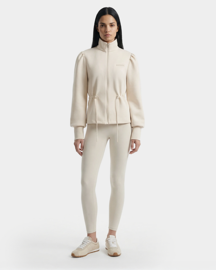 A woman with straight dark hair and a neutral expression wears the Rudsak ELODIE - 8526039 Iceland cream stretch zip-up sweater jacket with drawstrings, matching leggings, and white sneakers against a plain white background.