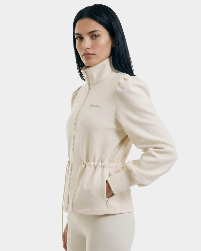 A woman with long dark hair wears the Rudsak ELODIE - 8526039 Iceland stretch cotton blend cream zip-up sweater jacket, featuring a high collar and drawstring waist. She stands against a plain light background, hands in her pockets.