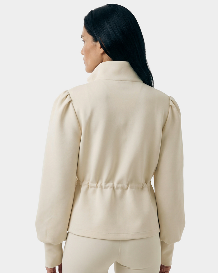 A woman with long black hair, seen from the back, wears the Rudsak ELODIE - 8526039 Iceland—an elegant beige zip-up sweater jacket with puffed sleeves and matching pants—against a plain light background.