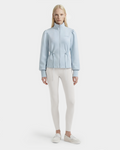 A woman with long blonde hair wears the Rudsak ELODIE - 8526039 Powder Blue zip-up sweater jacket, paired with white fitted leggings and white slip-on shoes, standing against a plain light background.