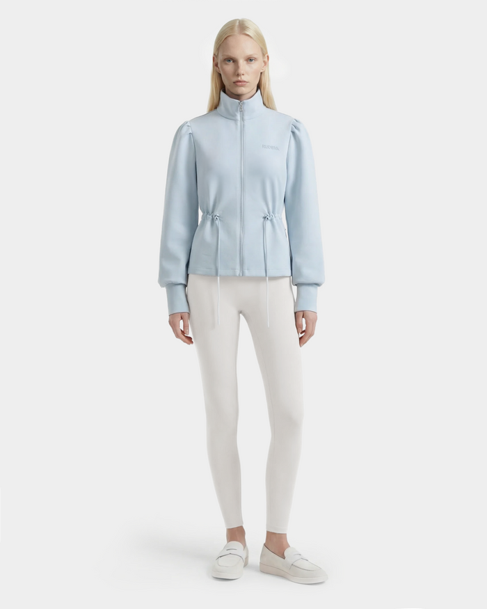 A woman with long blonde hair wears the Rudsak ELODIE - 8526039 Powder Blue zip-up sweater jacket, paired with white fitted leggings and white slip-on shoes, standing against a plain light background.