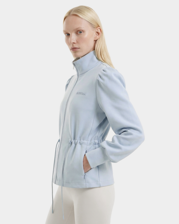 A woman with straight blonde hair wears the Rudsak ELODIE - 8526039 Powder Blue zip-up sweater jacket, featuring a high collar and drawstring waist, paired with white pants against a plain white background.