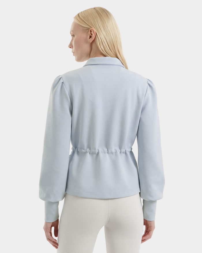 A woman with straight blonde hair is seen from the back wearing Rudsak’s ELODIE – 8526039 Powder Blue blouse and white pants, set against a plain white background.