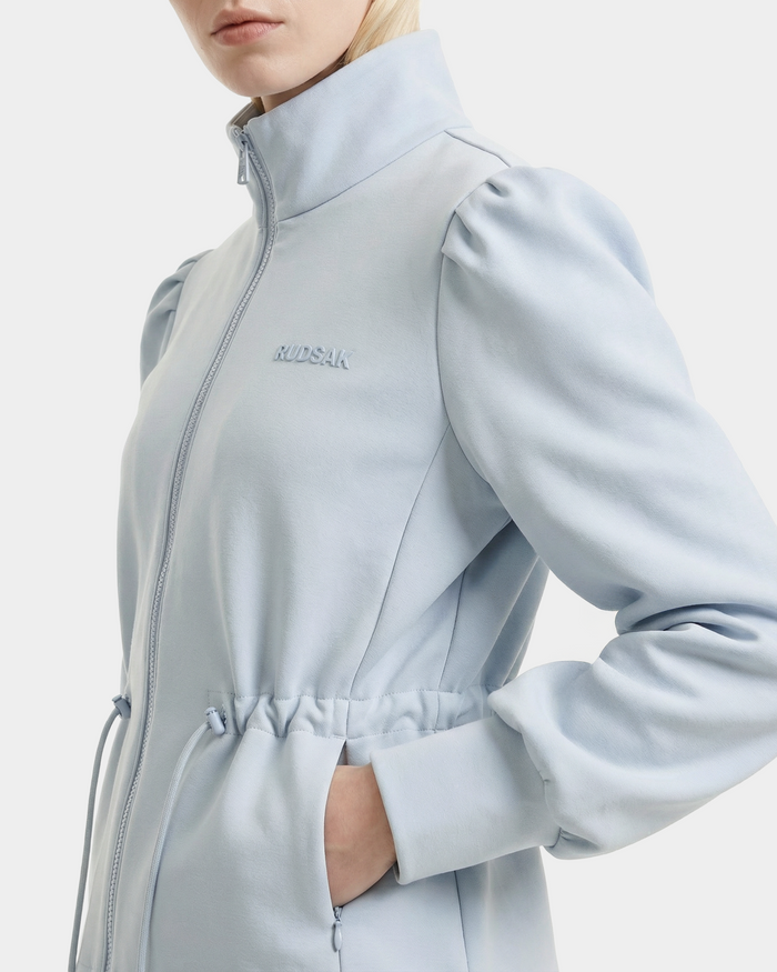 A person wears the Rudsak ELODIE - 8526039 Powder Blue zip-up sweater jacket, featuring puffed shoulders, chest embroidery, and a hand in the pocket. Made from a stretch cotton blend, it stands out against a plain white background.