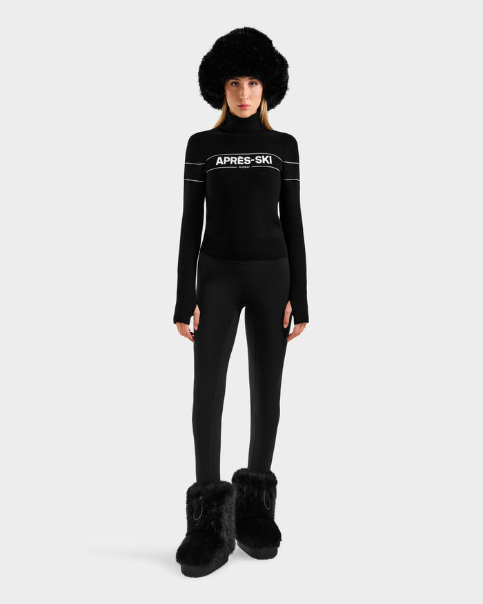 A person faces forward in a chic winter look, wearing the Rudsak ERIKA - 8523536 Black après-ski turtleneck sweater with black leggings, a large black fur hat, and furry boots against a plain light background.