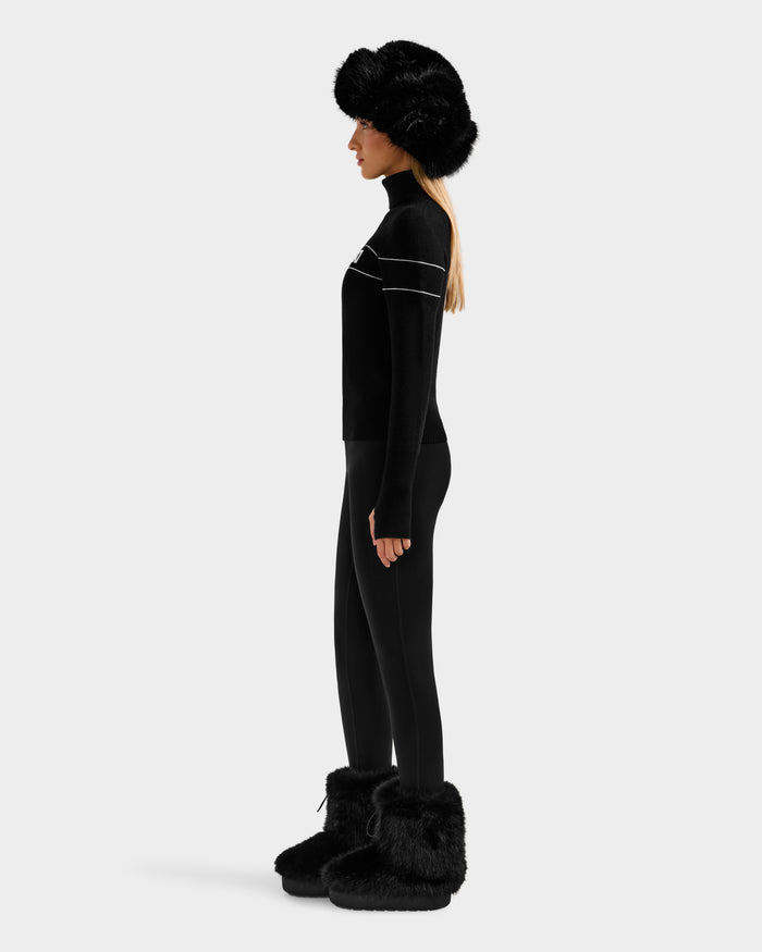 A person stands in profile wearing the Rudsak ERIKA - 8523536 Black long-sleeve top with white stripes, black leggings, a large black fur hat, and black fur boots—crafting a chic winter look on a light gray background.