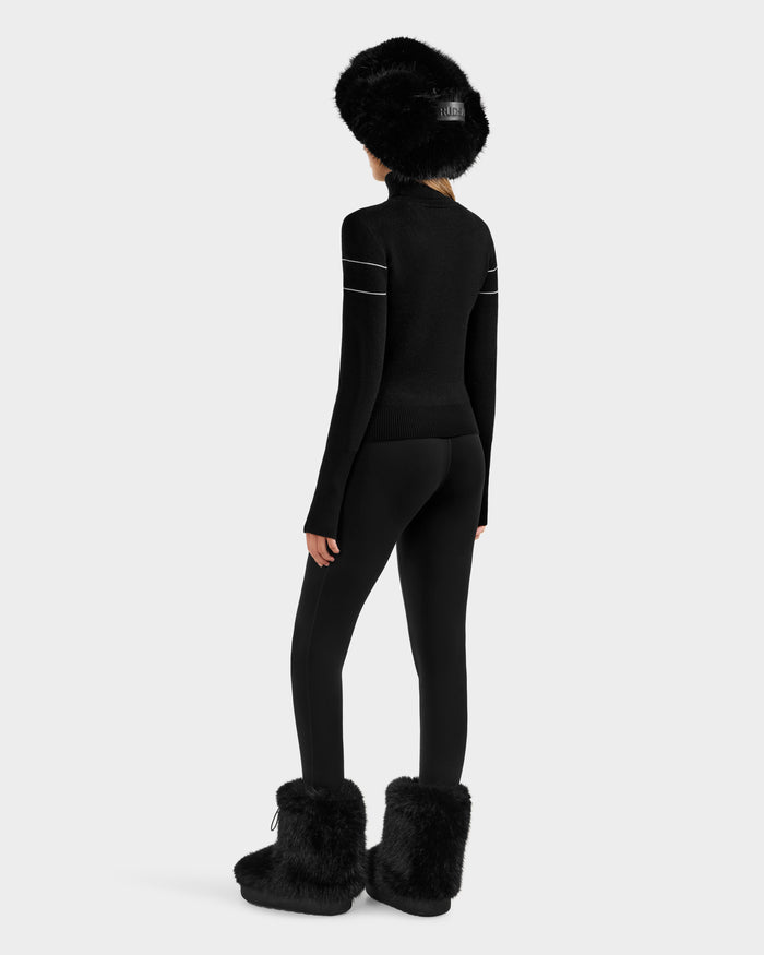 A person stands facing away, wearing the Rudsak ERIKA - 8523536 Black fur hat, a fitted black turtleneck sweater with white striped sleeves, black leggings, and black fur boots against a plain light background.