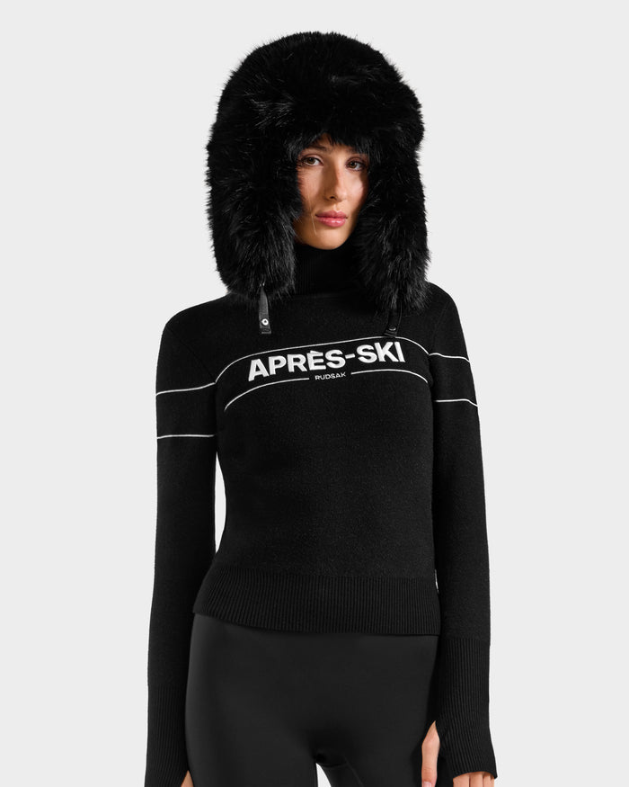 A person wears the Rudsak ERIKA - 8523536 Black, featuring a furry hood and APRÈS-SKI RUSLAN turtleneck, paired with black pants against a light gray background—showcasing a chic winter look.