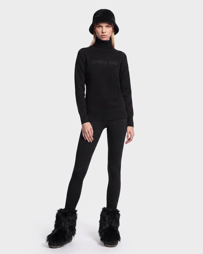 A person wearing the ERIKA - 8525705 Black turtleneck sweater by Rudsak, paired with a black bucket hat, black leggings, and black furry boots stands against a plain white background—a chic addition to any winter wardrobe.