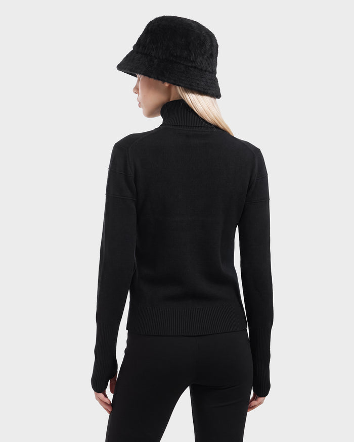 A woman stands facing away, dressed in the Rudsak ERIKA - 8525705 Black turtleneck sweater—a timeless staple for any winter wardrobe—paired with black pants and a black fuzzy bucket hat against a plain light gray background.