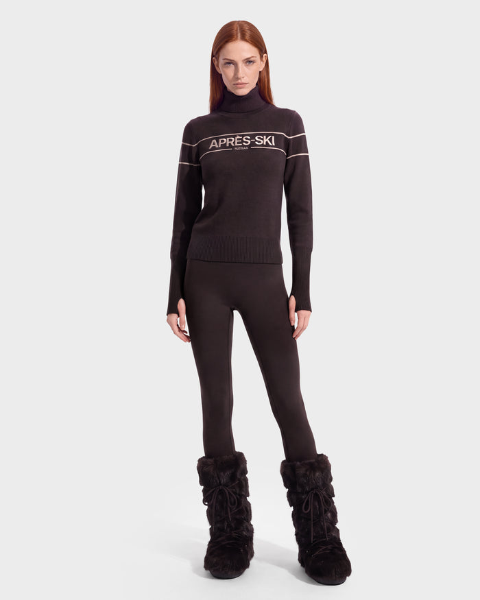 A woman wearing the ERIKA - 8525705 Espresso turtleneck sweater by Rudsak, paired with black leggings and black fur boots, stands against a plain white background, facing forward with a neutral expression—the perfect look to elevate any winter wardrobe.