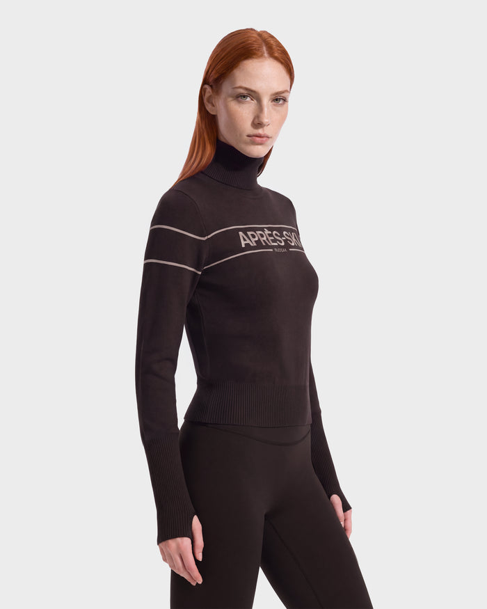 A woman with long red hair wears the Rudsak ERIKA - 8525705 Espresso fitted black après-ski turtleneck sweater with APRÈS-SKI text and matching black pants, standing against a plain white background—the perfect addition to a stylish winter wardrobe.