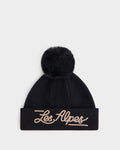 The Rudsak ESTELLA - 8426521 Black is a black knit beanie featuring a large pom-pom on top and “Les Alpes” embroidered in cursive beige letters on the folded brim, displayed against a plain white background.