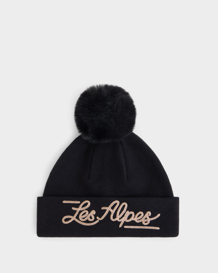 The Rudsak ESTELLA - 8426521 Black is a black knit beanie featuring a large pom-pom on top and “Les Alpes” embroidered in cursive beige letters on the folded brim, displayed against a plain white background.