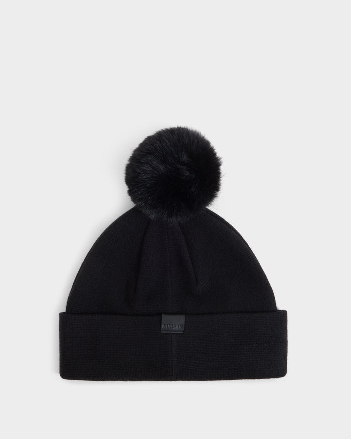 The Rudsak ESTELLA - 8426521 Black beanie features a large faux fur pom-pom on top and a folded cuff, shown against a plain white background.