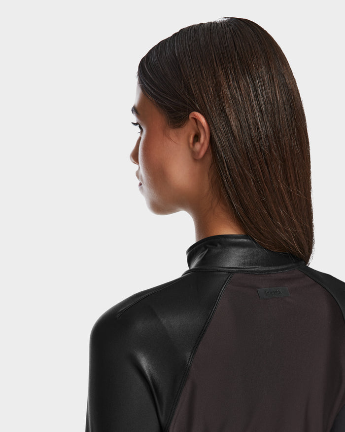 A woman with straight, wet-look dark hair is shown from behind and in profile, wearing the Rudsak EVA - 8524514 Black womens zip-up top with a high collar and mixed matte and shiny textures against a plain background.