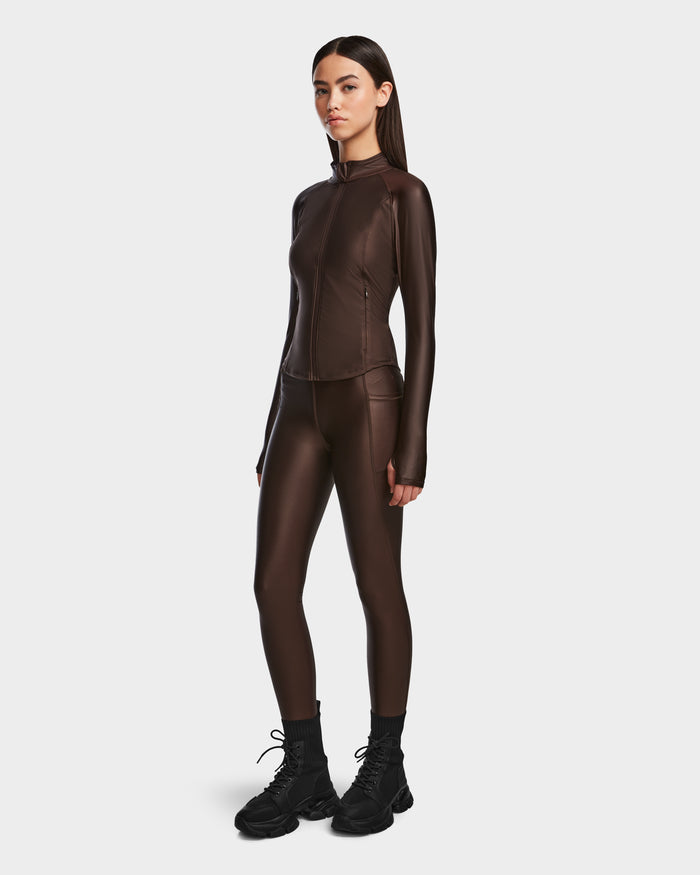 A woman wears the Rudsak EVA - 8524514 Espresso fitted zip-up top and matching leggings in shiny stretch fabric, paired with black lace-up boots. She has long, straight dark hair and poses against a plain light background.