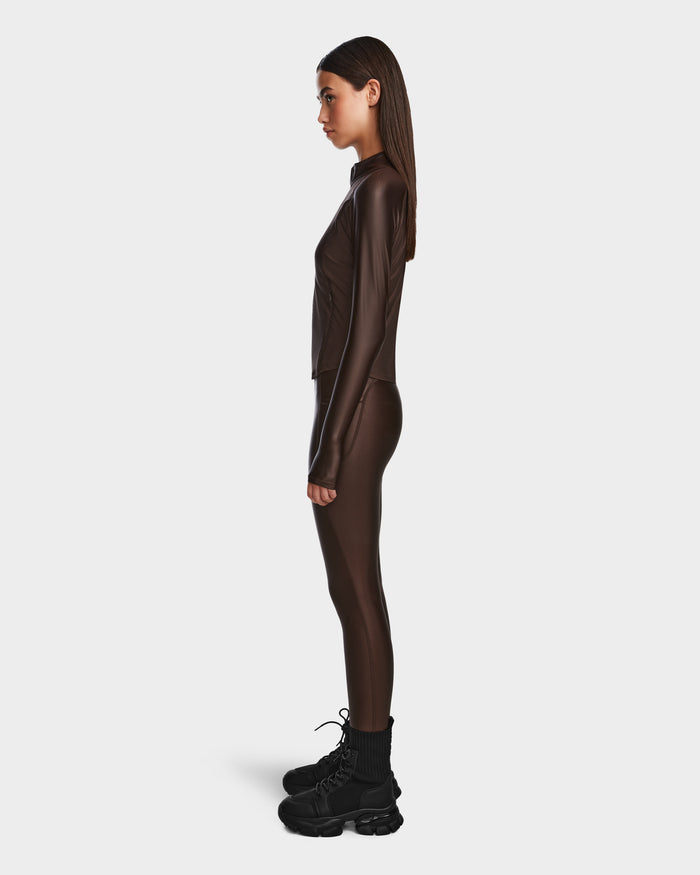 A woman stands in profile against a plain background, wearing the Rudsak EVA - 8524514 Espresso fitted zip-up top and matching leggings made from shiny stretch fabric, paired with black chunky boots.