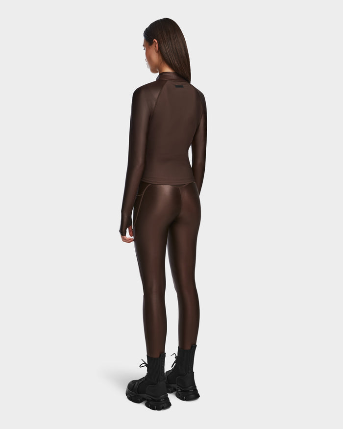 A woman faces away, wearing the Rudsak EVA - 8524514 Espresso fitted long-sleeve zip-up top with matching shiny stretch leggings and black chunky boots, set against a plain white background.