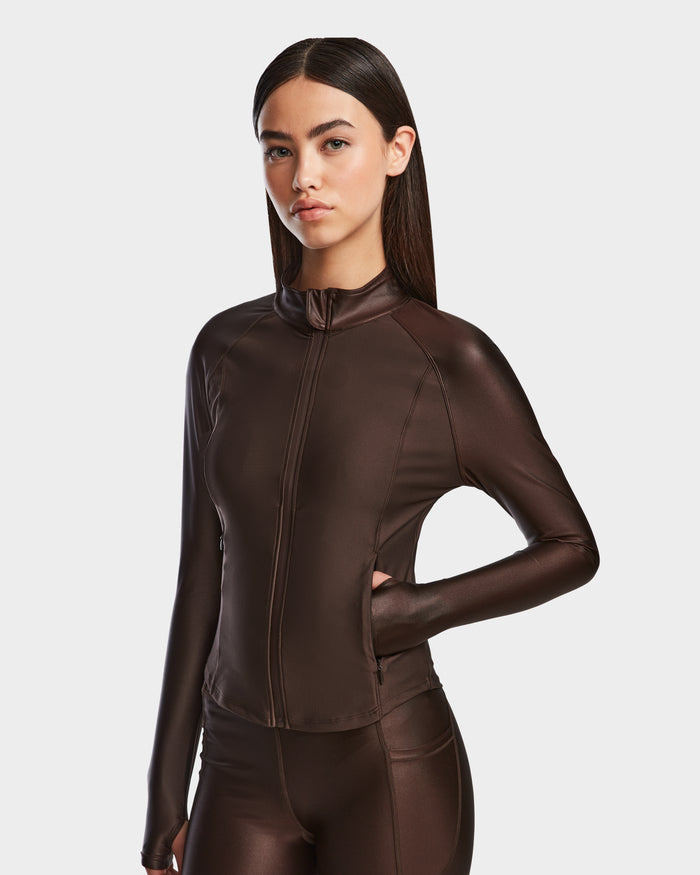 A woman poses with one hand on her hip, wearing the Rudsak EVA - 8524514 Espresso fitted zip-up top and matching pants in shiny stretch fabric with a waterproof zipper, set against a plain light background.