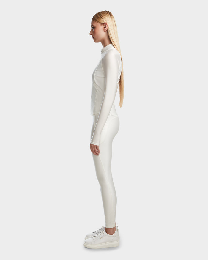 A woman with long straight blond hair stands in profile wearing the Rudsak EVA - 8524514 Iceland fitted zip-up top and matching white leggings, both in shiny stretch fabric, paired with white platform sneakers against a plain light background.