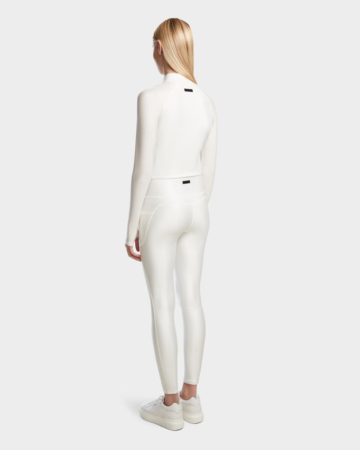 A person with long blonde hair faces away, wearing the Rudsak EVA - 8524514 Iceland fitted womens zip-up top with a water-proof zipper and matching shiny white leggings, paired with white sneakers against a plain light background.