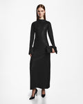 A woman stands against a white background wearing the Rudsak FAIZA - 8524579 Black maxi dress with black heels and a small black clutch. Her straight, long brown hair completes the sophisticated ensemble.