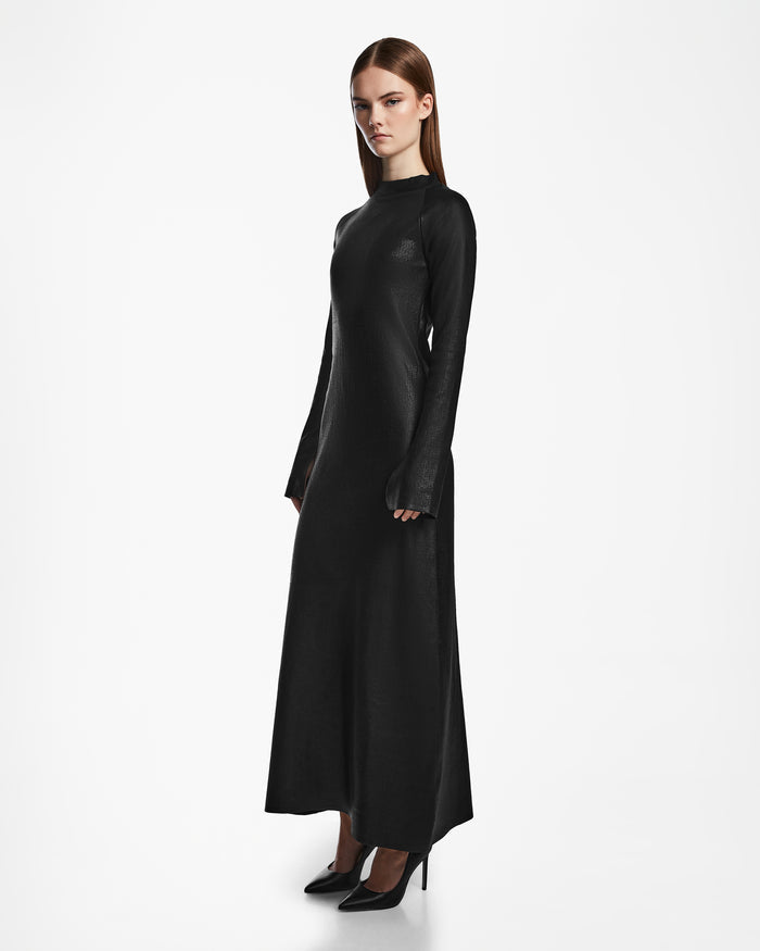 A woman with straight brown hair stands on a white background, wearing the Rudsak FAIZA - 8524579 Black mock-neck long sleeve dress and black high heels. She faces slightly to the side with a neutral expression.