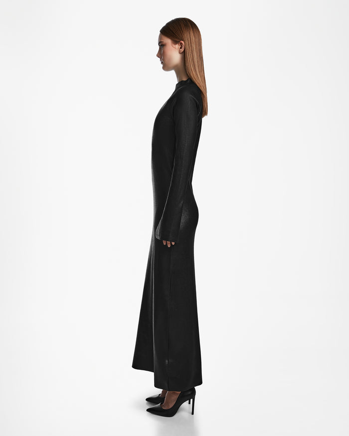 A woman with straight, long hair stands in profile wearing the Rudsak FAIZA - 8524579 black, long-sleeved, floor-length mock-neck dress and black high heels against a plain white background.