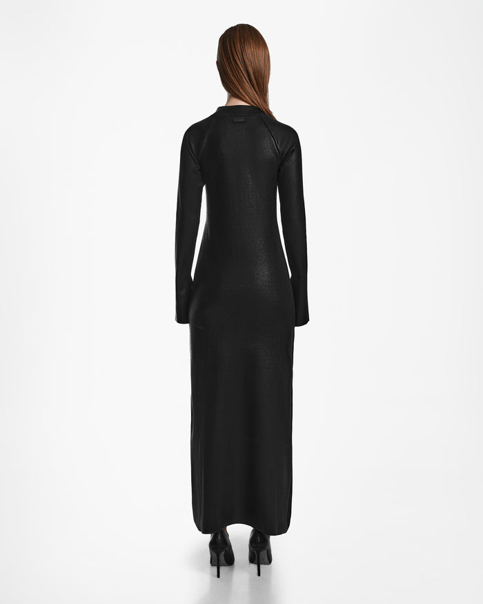A woman with long brown hair, wearing the Rudsak FAIZA - 8524579 Black polished-finish maxi mock-neck dress and black high heels, stands facing away from the camera against a plain white background.