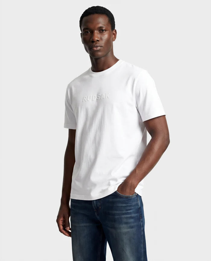 A man wears the Rudsak FERDI - 6522005 NATURAL WHITE T-shirt with subtle chest branding and blue jeans, standing against a light gray background, one hand in his pocket.