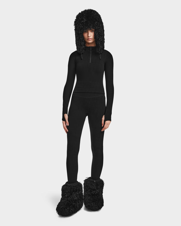 Dressed head-to-toe in Rudsak’s FERKA - 8524542 Monogram Print Black, featuring a long-sleeve top, thermal leggings, and an oversized furry hat with ear flaps and matching slippers—ideal for a cozy, stylish winter look.