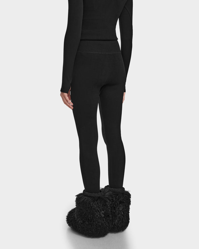 A person stands with their back to the camera, wearing Rudsaks FERKA - 8524542 Monogram Print Black leggings and a fitted black top, paired with large, fluffy slippers—ideal for adding style to your winter wardrobe against a plain white background.