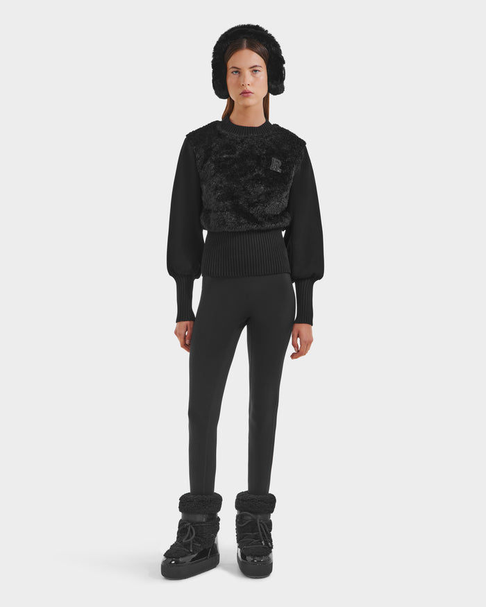 A woman faces forward wearing the FLORENCE - 8525546 Black hat by Rudsak, along with a textured top, fitted black pants, and fur-accented boots—an après-ski must-have—set against a plain white background.