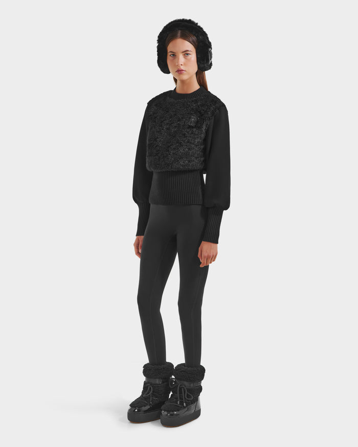 A woman with straight brown hair and a neutral expression models the Rudsak FLORENCE - 8525546 Black earmuffs against a plain background, paired with a textured black top, black leggings, and winter boots—an après-ski essential.
