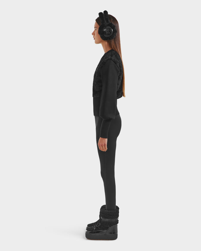 A woman in profile poses against a plain background wearing the Rudsak FLORENCE - 8525546 Black: an all-black après-ski look with faux fur earmuffs featuring bunny ears, a textured top, leggings, winter boots, and her long hair straight and loose.