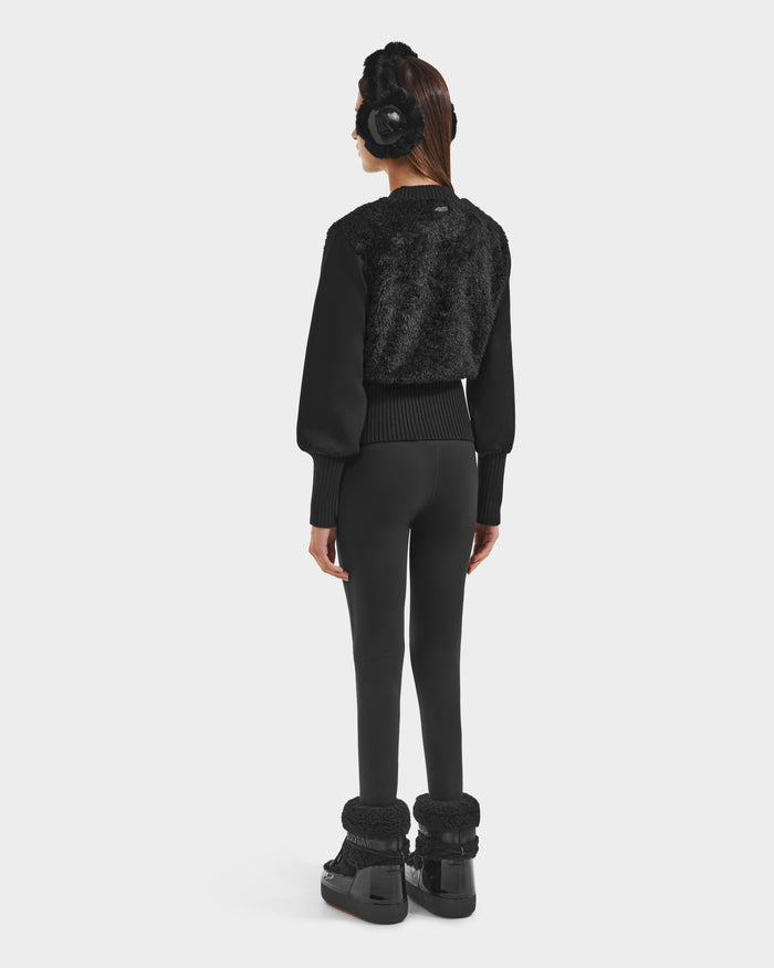 A woman stands facing away, wearing Rudsaks FLORENCE - 8525546 Black winter boots with fur trim—an après-ski essential—paired with black earmuffs, a faux fur cropped womens top, and leggings against a plain white background.