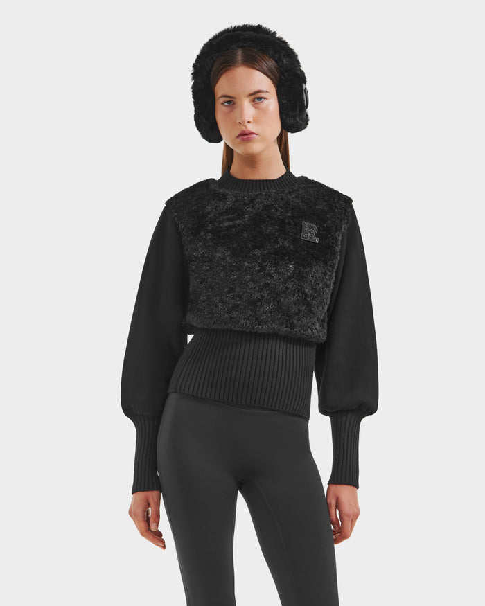 A woman wears the Rudsak FLORENCE - 8525546 Black faux fur hat, a textured après-ski cropped top with long sleeves, and black fitted pants, standing against a plain white background.