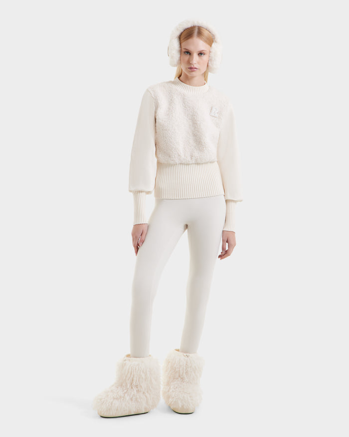 A woman stands against a plain background, exuding cozy glamour in Rudsaks FLORENCE - 8525546 Iceland boots, fluffy earmuffs, a textured cream sweater, and beige leggings—a chic, monochromatic winter look with après-ski appeal.