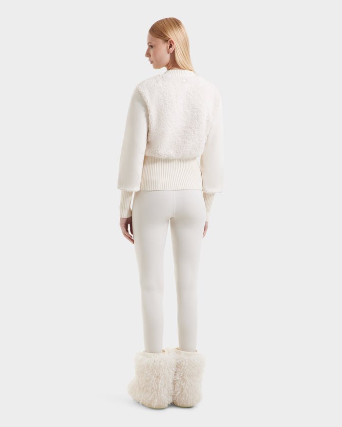 A woman with straight blonde hair stands facing away, exuding cozy glamour in the Rudsak FLORENCE - 8525546 Iceland white textured jacket, fitted white pants, and large fluffy après-ski boots against a plain light background.