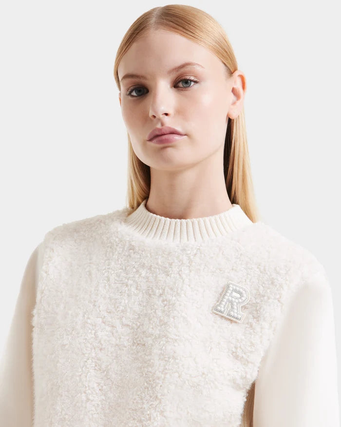 A woman with straight blonde hair wears the Rudsak FLORENCE - 8525546 Iceland cream textured sweater with a ribbed collar and decorative letter R pin, exuding cozy glamour as she looks slightly to the side against a plain light background.