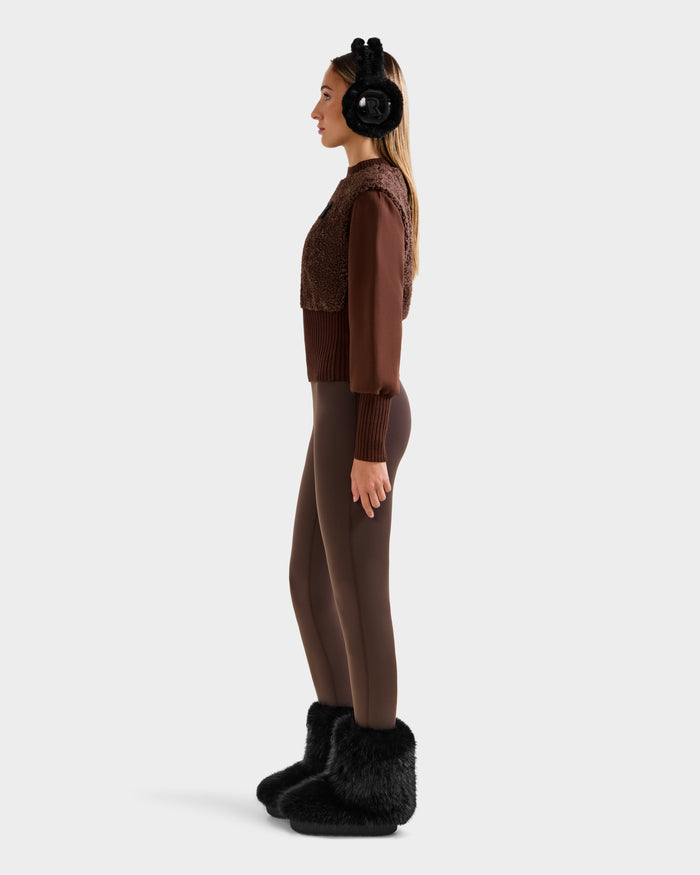A woman in profile wears Rudsak’s FLORENCE - 8525546 Mocha faux fur top as an après-ski essential, styled with black fuzzy earmuffs, brown leggings, and black furry slippers against a plain light background.
