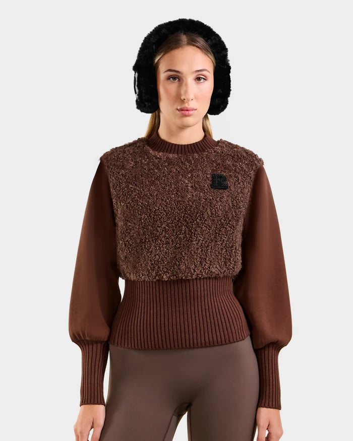 A woman stands facing forward against a plain light background, exuding cozy glamour in the Rudsak FLORENCE - 8525546 Mocha textured après-ski top with ribbed cuffs and hem, brown leggings, and black faux fur earmuffs.