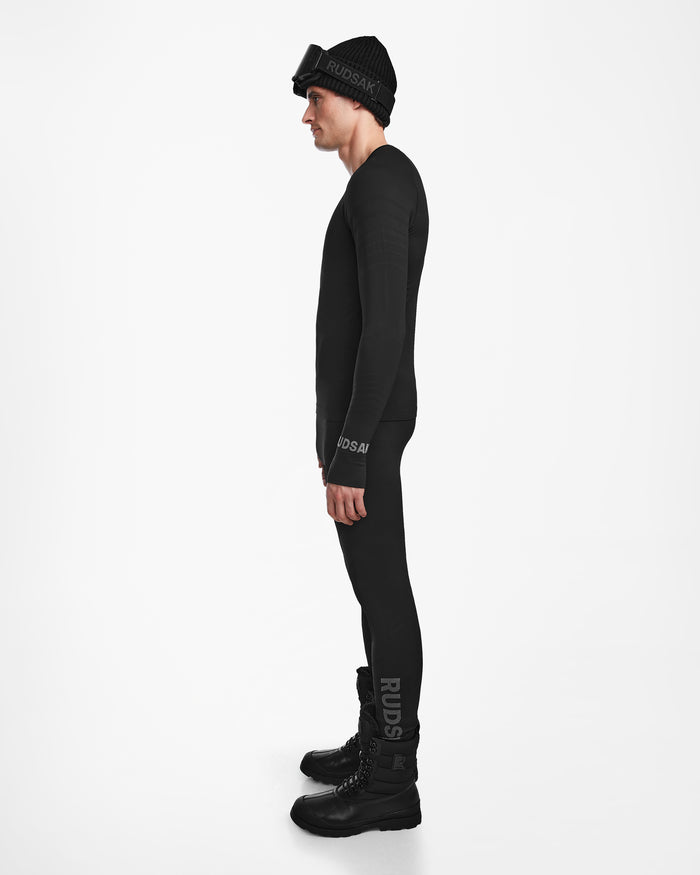 A man in profile wears the Rudsak FRANC - 6524541 Black men’s thermal base layer set—leggings and a long-sleeve top—paired with boots, a beanie, and ski goggles, ideal as a skiing underlayer against a plain white background.
