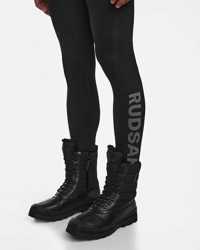 A model wears Rudsak FRANC - 6524541 Black leggings, perfect as a ski underlayer, with large gray “RUDSAK” lettering on one leg, paired with Rudsak black lace-up winter boots featuring padded sides and rugged soles. Background is plain white.