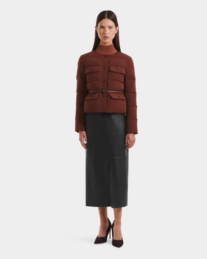A woman faces forward in the Rudsak FRANCESCA - 8125021 Rust fitted, dark red quilted puffer jacket with front pockets and a vegan leather belt, paired with a long black leather skirt and heels, set against a white background.
