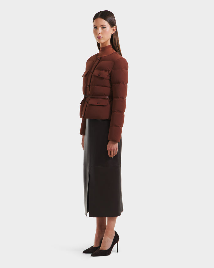A woman stands in profile against a white background wearing the Rudsak FRANCESCA - 8125021 Rust fitted brown quilted puffer jacket, styled with a black midi skirt, black high heels, and a vegan leather belt.