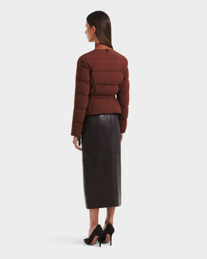 A woman faces away in the Rudsak FRANCESCA - 8125021 Rust puffer jacket, paired with a black midi leather skirt and black high heels, set against a plain white background.